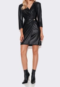 Donna che indossa un vestito avvolgente in ecopelle nera con maniche a tre quarti e stivaletti neri con tacco a spillo.