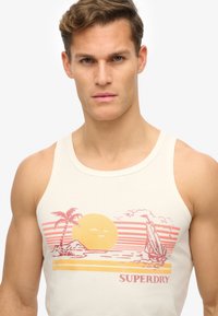 Top de tirantes beige con un gráfico de escena playera, que presenta un sol, palmeras y un velero, acentuado por rayas horizontales rojas y amarillas.