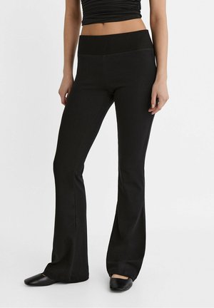 Frau, die hoch taillierte schwarze Schlaghosen mit einem gerippten Bund und schwarzen Slippern trägt, steht vor einem weißen Hintergrund.