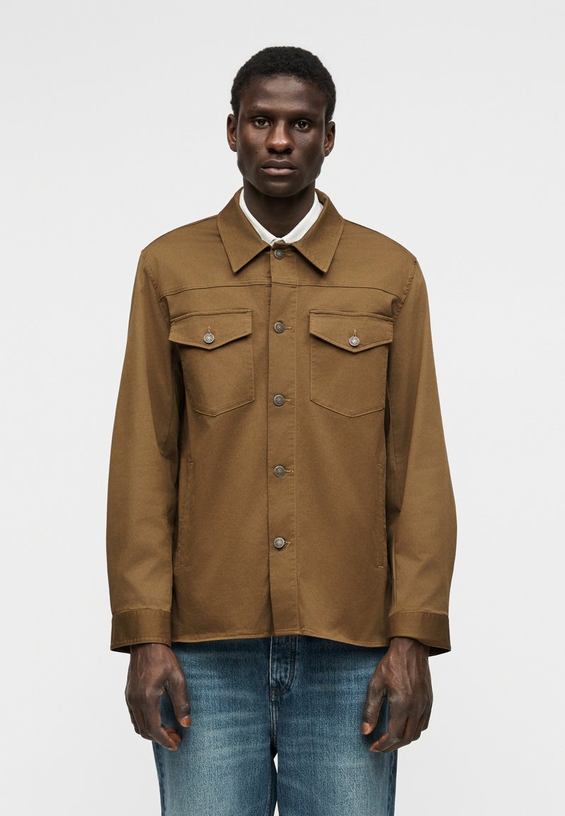 Mann trägt eine braune, knöpfbare Jacke mit Brusttaschen über einem weißen Hemd mit Kragen, kombiniert mit blauen Jeans, stehend vor einem schlichten Hintergrund.