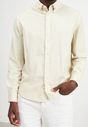 Mężczyzna w kremowej koszuli na guziki z długim rękawem, z jedną ręką w kieszeni, stojący na tle jednolitego jasnego tła.