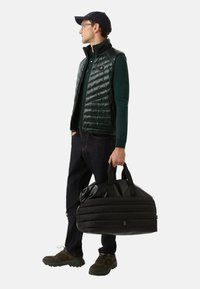 Mann trägt eine dunkelgrüne Mütze, Brille, grüne Langarmbluse, schwarze gepolsterte Weste und Schuhe, hält eine schwarze gepolsterte Sporttasche und steht seitlich.