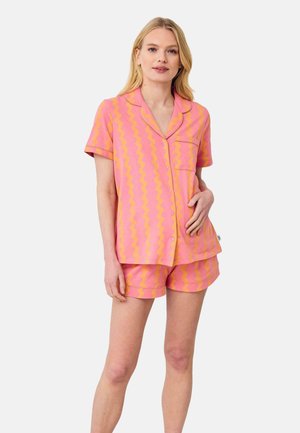 Kvinde stående iført matchende kortærmet skjorte med knapper og shorts med pink og orange bølget stribemønster på ensfarvet baggrund.