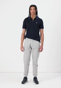 Polo bleu marine à manches courtes et col zippé, associé à un pantalon de survêtement gris clair et des chaussures de sport noires. Présente des accents bleus.