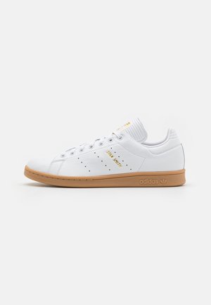 Baskets basses blanches Adidas Stan Smith avec semelle en gomme, lettrage doré et bandes latérales perforées sur fond blanc uni.