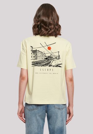 Yellow cotton t-shirt featuring a black line drawing of a street scene with a red sun. Text below reads "ESCAPE AND DISCOVER THE WORLD."