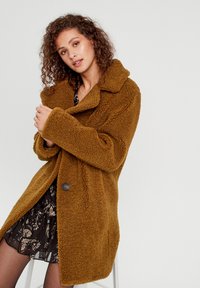 Manteau marron, duveteux et surdimensionné avec un grand col et une fermeture à bouton. Porté par-dessus une robe foncée à motifs, mettant en valeur un tissu texturé.