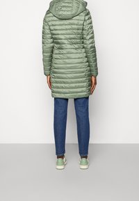 Lys grøn polstret jakke med hætte, der har vandret kviltet design. Bæres over blå jeans og grønne sneakers.