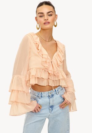 Femme portant un crop top long à manches longues couleur pêche avec des volants et un nouage à l'avant, associée à un jean taille haute bleu clair, posant les mains dans les poches.