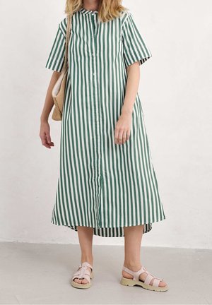 Femme portant une robe midi à rayures verticales vertes et blanches avec des manches courtes, des sandales beiges, et portant un sac à bandoulière en osier beige.