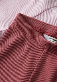 Primo piano di abbigliamento in tessuto costellato color rosa polveroso con cuciture visibili e etichetta parziale che mostra la misura 2-3 mesi.