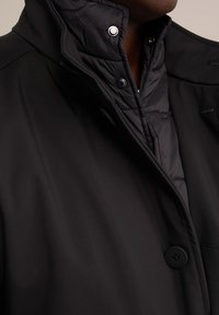 Abrigo negro con un cuello interior acolchado, botones a presión y una tela suave y texturizada. Detalle en primer plano del cuello y el botón.