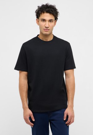 Göndör hajú férfi, egyszínű fekete rövid ujjú pólót és sötétkék nadrágot visel, világosszürke háttér előtt áll.