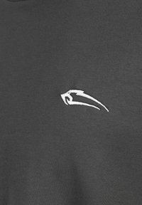 Camiseta gris oscura con una textura de tela suave. Presenta un pequeño logo bordado en blanco en la parte delantera, que representa un animal estilizado.