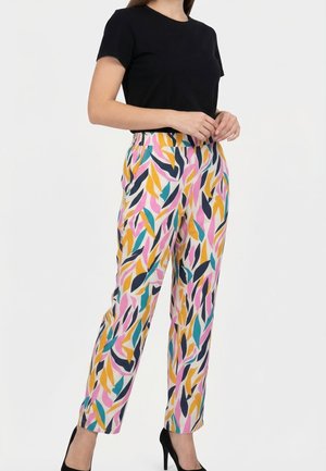 Donna che indossa una maglia nera a maniche corte e pantaloni dritti con fantasia colorata a foglie, con tacchi alti neri, in piedi davanti a uno sfondo bianco.