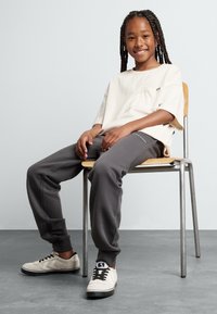 Beige rövid ujjú póló zsebbel, szürke tréningnadrág kiegészítővel, és krém színű sneaker; egy fém székben ülve, világos háttér előtt.