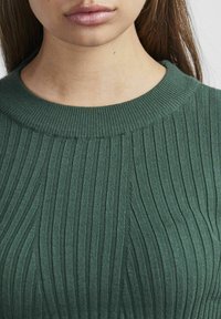 Femme portant un pull en maille côtelée vert avec un col rond, montrant le bas du visage, le cou et le haut de la poitrine.