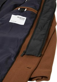 Brun ytterlag med en marineblå innside, med glidelåslukking og lommer. Merket "SELECTED HOMME" for ansvarlig produksjon.