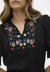 Svart klänning med en knappslå som har färgglad blommönstring i röd, rosa, gul och blå. Mjuk, texturerad tyg och justerbara band.