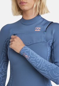 Combinaison de plongée bleue avec une poche zippée sur la poitrine. Caractéristiques : motifs abstraits sur les manches, coutures plates et un accent de logo en corail.