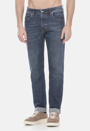 Hombre con vaqueros azules remangados y zapatillas taupe de pie contra un fondo blanco.