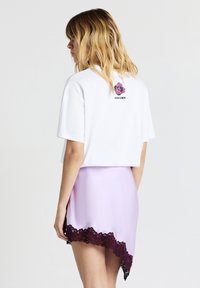 Donna con capelli biondi fino alle spalle, indossa una maglietta corta bianca con una grafica di fiori e una gonna lilla chiara con bordi in pizzo scuro.