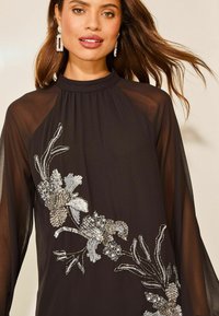 Rochie neagră cu mâneci lungi din material transparent și guler înalt. Decupată cu mărgele argintii intricate în modele florale pe o parte.