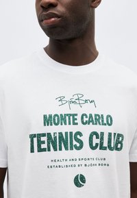 Άνδρας που φοράει λευκό μπλουζάκι με πράσινο κείμενο "Björn Borg Monte Carlo Tennis Club Health and Sports Club Established by Björn Borg" και εικονίδιο μπάλας του τένις.