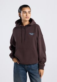 Sudadera grande marrón con un bolsillo frontal, capucha con cordón y texto bordado en azul claro que dice "Hotel de Nueva York". Material de algodón.