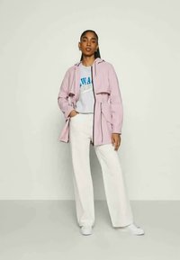 Casaco capuz rosa claro com faixa na cintura, usado sobre um sweatshirt cinza e calças brancas de perna larga, combinado com tênis brancos.