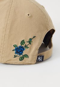 Baseballkappe aus beigem Stoff mit blauer Rosenstickerei und grünen Blättern. Ausgestattet mit einem verstellbaren Riemen und einem Logo-Anhänger an der Seite.