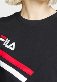 Gros plan d'un sweat-shirt noir FILA avec un logo brodé et des rayures rouges et blanches sur la poitrine, porté par une personne aux cheveux blonds courts.