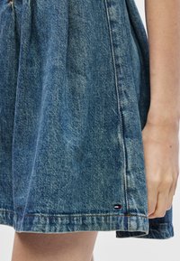 Denim φόρεμα σε μεσαία μπλε απόχρωση με μαζεμένη μέση, διαθέτοντας ένα στρίφωμα με αντί�θετη ραφή και μια μικρή λογότυπη λεπτομέρεια στη πλευρά.