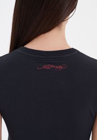 Sort bomulds T-shirt med rund halsudskæring og korte ærmer. Den har rød broderet "Ed Hardy" tekst på den øverste ryg.