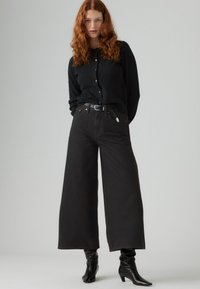 Svarte vide jeans kombinert med en svart knapp-up cardigan. Merkbare detaljer inkluderer et belte med sølvspenne og spisse svarte ankelstøvler.