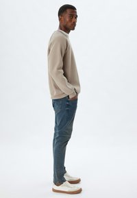 Sudadera beige clara con puños acanalados, combinada con jeans azules y zapatillas blancas con acentos marrones. Mostrada en un perfil lateral.