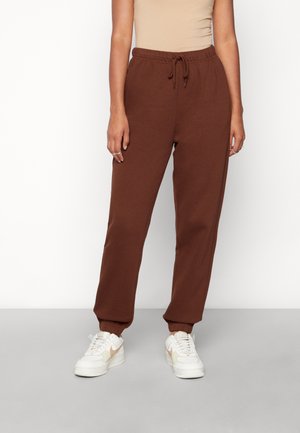 Personne portant un pantalon de jogging marron avec cordon et des baskets blanches, debout devant un fond blanc uni.