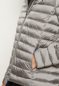 Veste matelassée grise avec un design matelassé horizontal, fermeture éclair et poches latérales. Elle présente une texture lisse et brillante ainsi qu'une silhouette moderne.