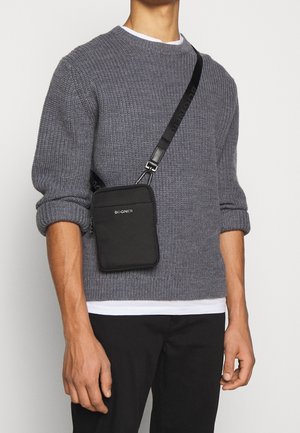 Homme portant un pull en maille gris et un sac bandoulière noir avec une large sangle portant l'inscription "BOGNER" par-dessus une chemise blanche et un pantalon noir.