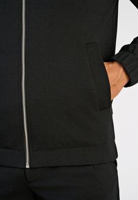 Zwarte jas met ritssluiting, voorzien van een gladde textuur, zijzakken en elastische manchetten. De ritssluiting is zilverkleurig met een strak, minimalistisch ontwerp.