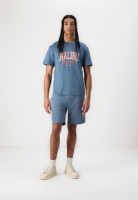 Blauwe katoenen t-shirt met "MALIBU" grafiek, gecombineerd met bijpassende shorts. Crème sneakers en lichte sokken maken de casual look compleet.