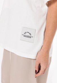 Hvit bomulls t-skjorte med et firkantet brodert merke som har teksten "All we got THE LITTLE THINGS IN LIFE PEGADOR" i svart.