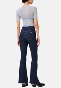 Camicia azzurro chiaro, aderente, con maniche corte; jeans in denim blu scuro a palazzo con tasche posteriori e un logo discreto sulla cintura. Calzature con tacco nere.