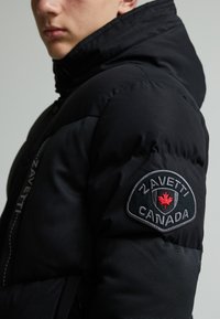 Doudoune noire avec capuche amovible, poches latérales et un patch brodé avec "Zavetti Canada" et une feuille d'érable rouge.