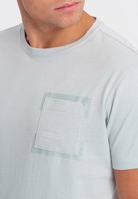 T-shirt in cotone grigio chiaro con taschino sul petto e una toppa a contrasto. Collo a costine e tessuto morbido, adatto per l'abbigliamento casual.