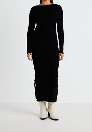 Czarna, prążkowana sukienka z długim rękawem, okrągłym dekoltem oraz bocznym rozcięciem, wykonana z teksturowanej tkaniny; zestawiona z jasnymi botkami na obcasie.