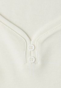 Cream-colored cotton shirt featuring a V-neck and two white buttons at the neckline, with a soft, ribbed texture.