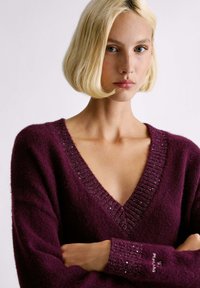 Maglione viola a maglia con un profondo scollo a V, rifinitura a coste testurizzata e sottili accenti brillanti sulle maniche. Materiale morbido e accogliente.