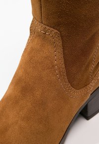 Gros plan sur une botte en daim marron avec des coutures détaillées et un talon empilé noir sur un fond blanc.