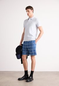 Ανοιχτό γκρι πόλο μπλούζα με διακριτό λογότυπο, σε συνδυασμό με μπλε καρό σορτς και μαύρες loafers. Το μοντέλο κρατά μια μαύρη ζακέτα και μια μπλε τσάντα.